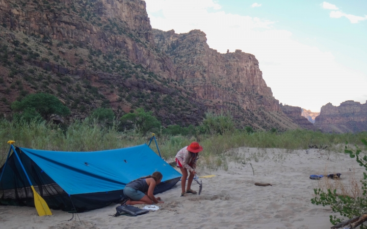 Two people set up a tarp shelter in the sand. In the background, tall canyon walls reach toward a blue sky.
